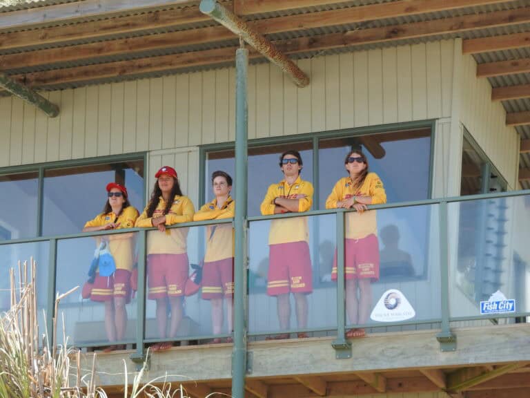 Raglan Surf Lifesaving team members standing on the clubhouse balcony, wearing yellow uniforms, ready for surf rescue duties at Raglan Beach, New Zealand.
