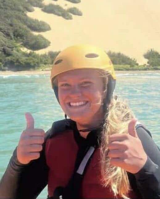 1. Female surf lifesaver smiling in yellow helmet and wetsuit giving thumbs up at Raglan Beach, demonstrating surf lifesaving skills and safety.